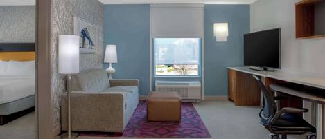 A hotel room with a bed, a sofa, a desk with a TV, a window with a view, and a patterned rug.