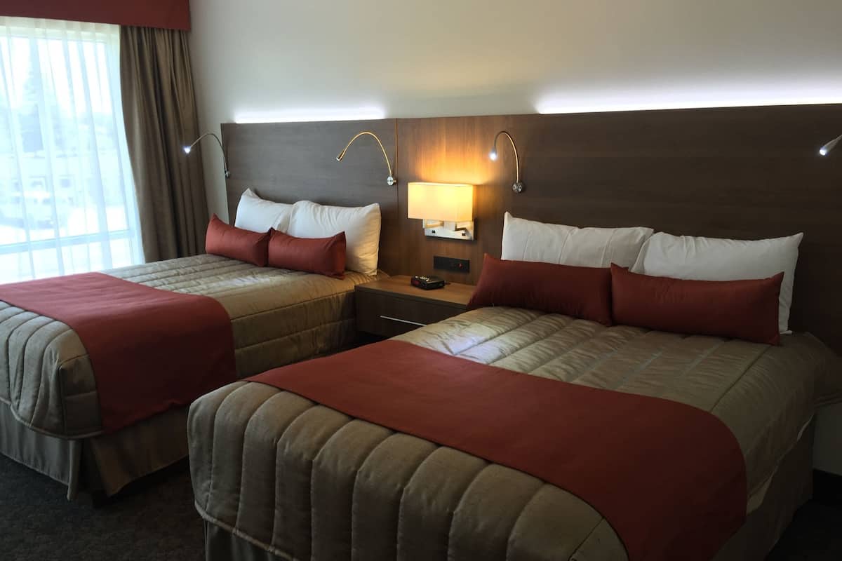 A hotel room with two beds, each with a headboard, a nightstand with a lamp, and a window with curtains.