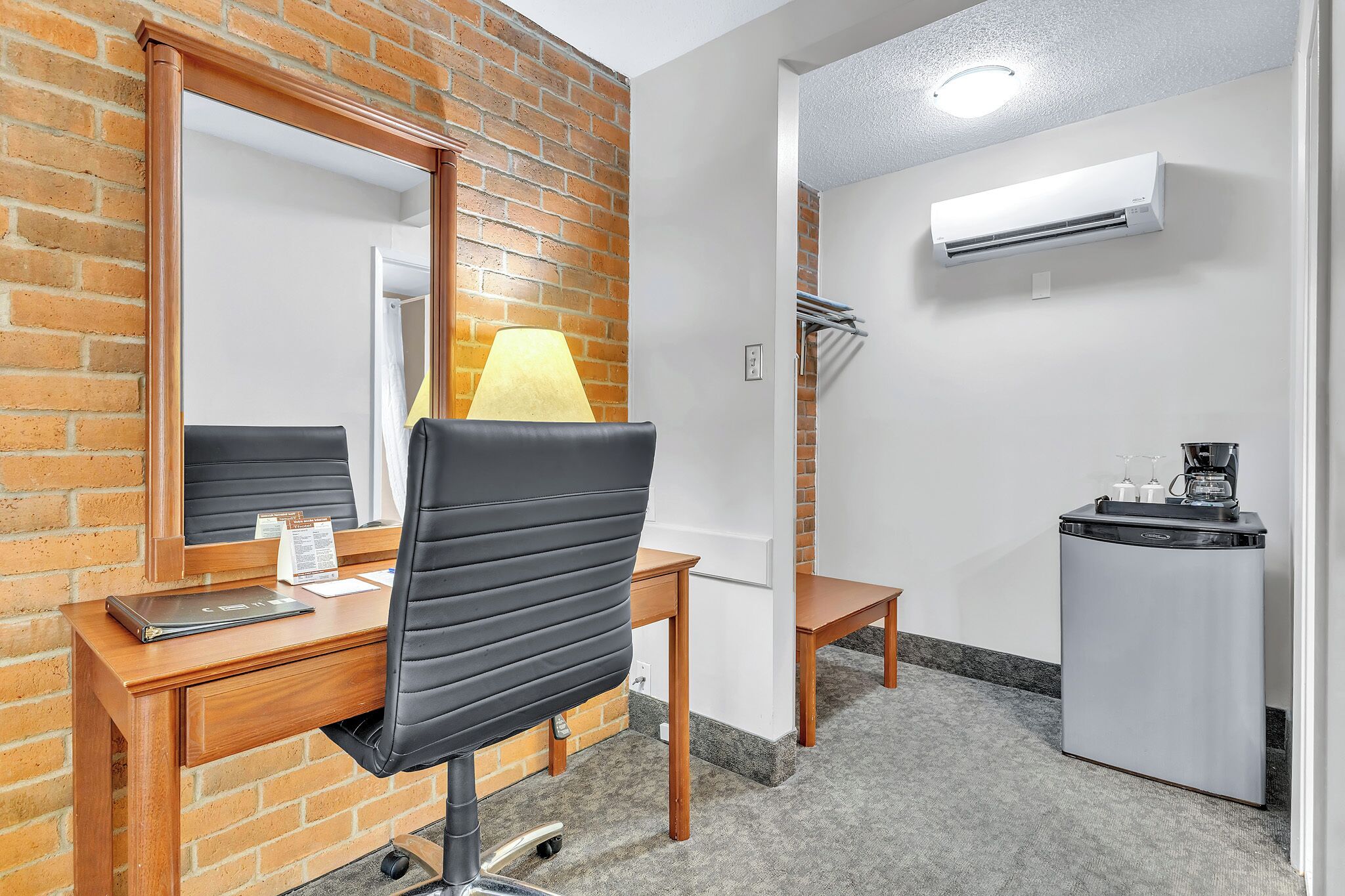Habitación de hotel con escritorio de madera, una silla de oficina negra, espejo, lámpara, mesita pequeña, refrigerador y aire acondicionado.