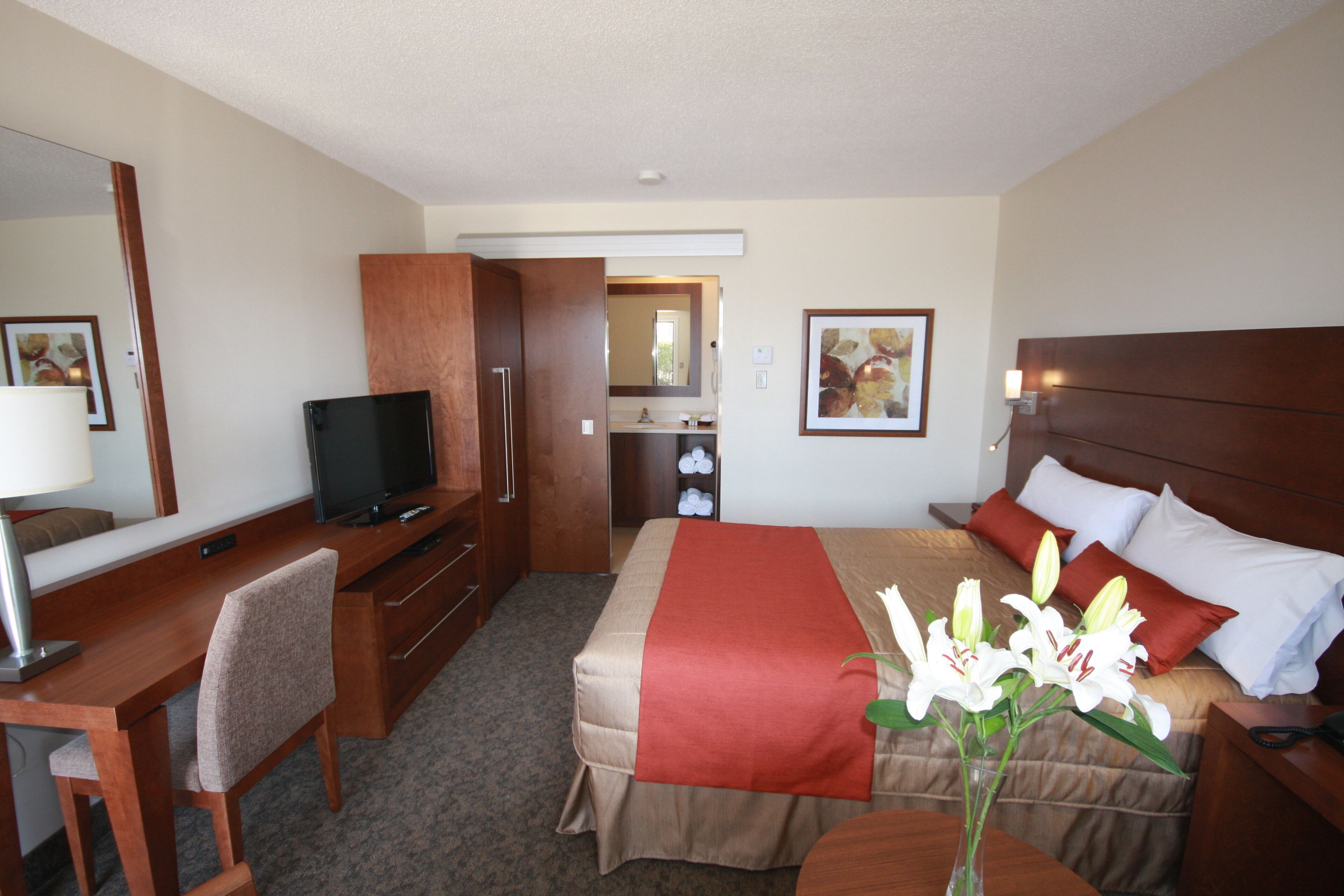 Une chambre d’hôtel comprenant un lit, un bureau, une chaise, une télévision, un miroir et un vase avec des fleurs.