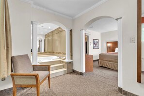 Een hotelkamer met een groot bed, een zithoek met een stoel, een badkamer met een bad, en een hal met een spiegel.