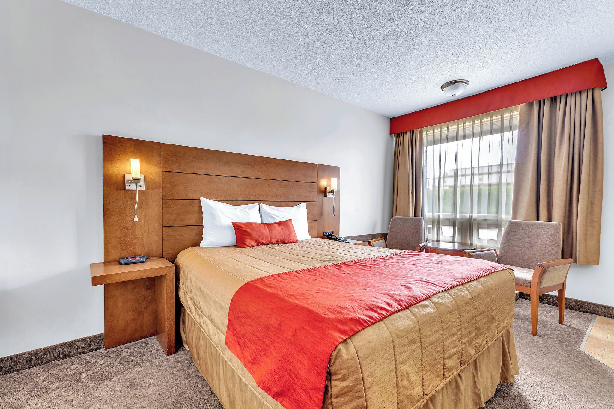 Habitación de hotel con cabecera de madera, cama con ropa de cama roja, mesita de noche con teléfono, una silla y una ventana con cortinas.