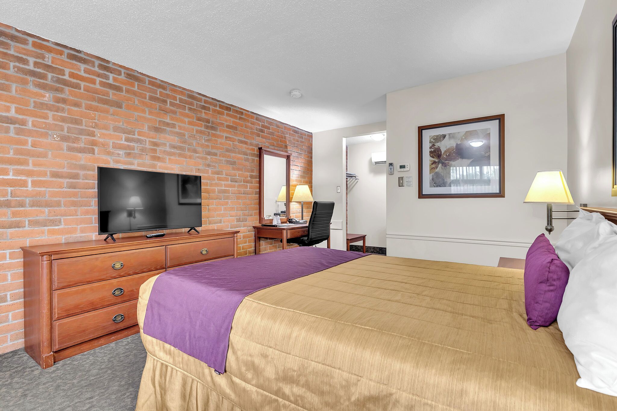 Quarto de hotel com parede de tijolos, televisão, escrivaninha com abajur, cama com travesseiro roxo e branco e um quadro pendurado na parede.