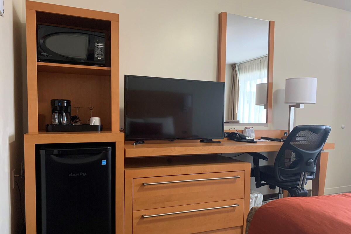 A hotel room with a wooden desk, a television, a coffee maker, a refrigerator, a chair, and a bed.
