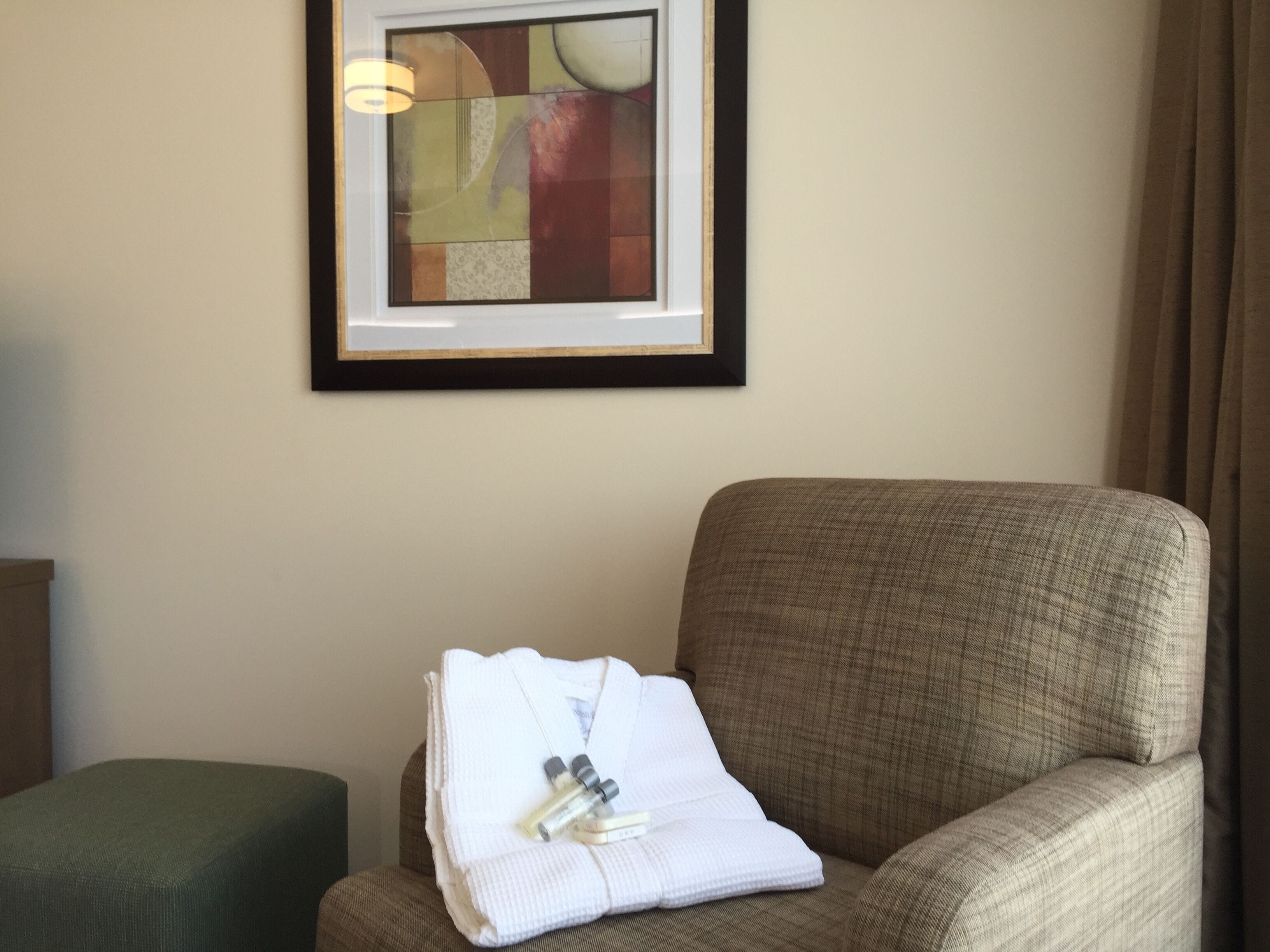 Habitación de hotel con un cuadro abstracto enmarcado en la pared, un reposapiés verde y una silla con una toalla blanca doblada y artículos de tocador.