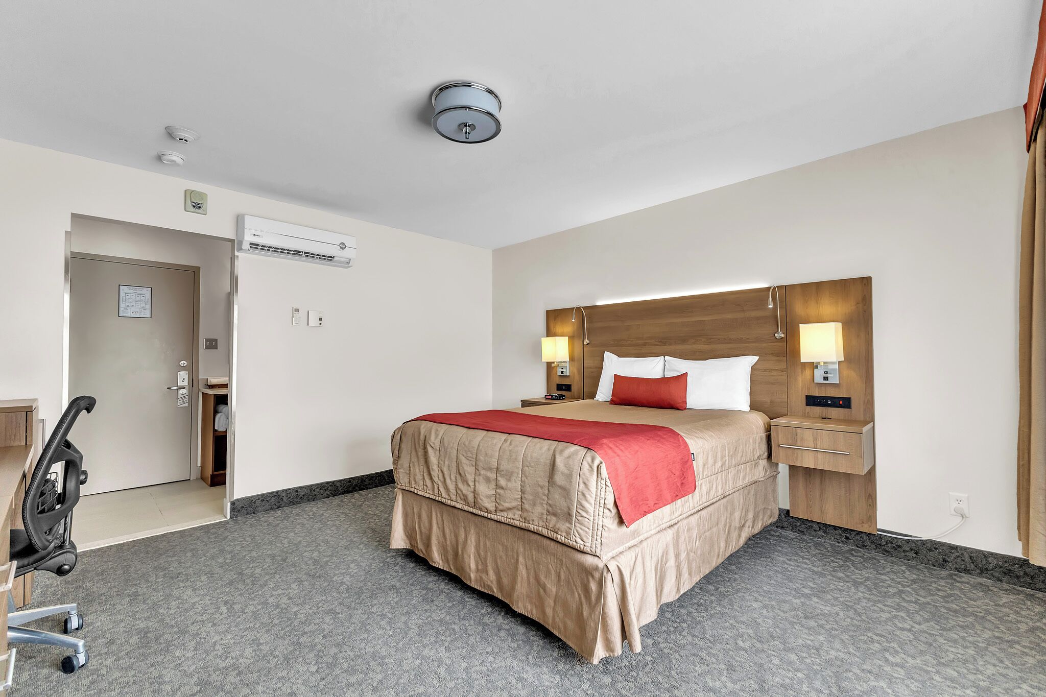 Een hotelkamer met een bed, nachtlampjes, een airconditioning en een deur, plus een rolstoel.