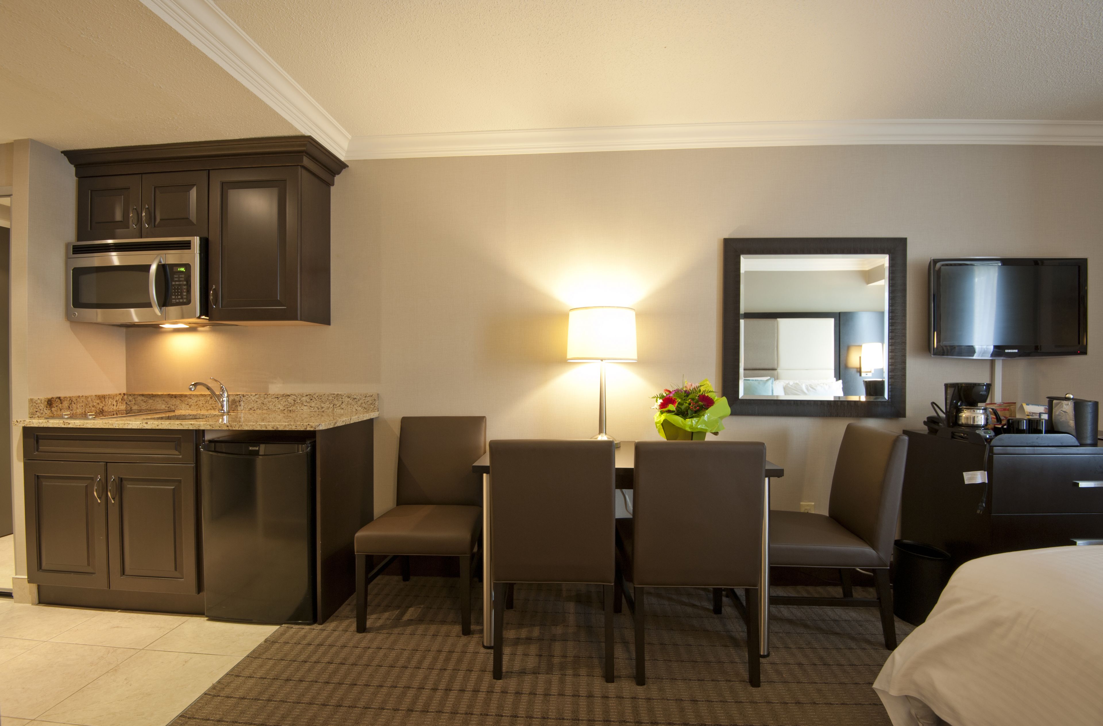 A hotel room with a kitchenette, a dining area with chairs and a table, a lamp, a mirror, and a TV.
