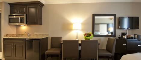 A hotel room with a kitchenette, a dining area with chairs and a table, a lamp, a mirror, and a TV.