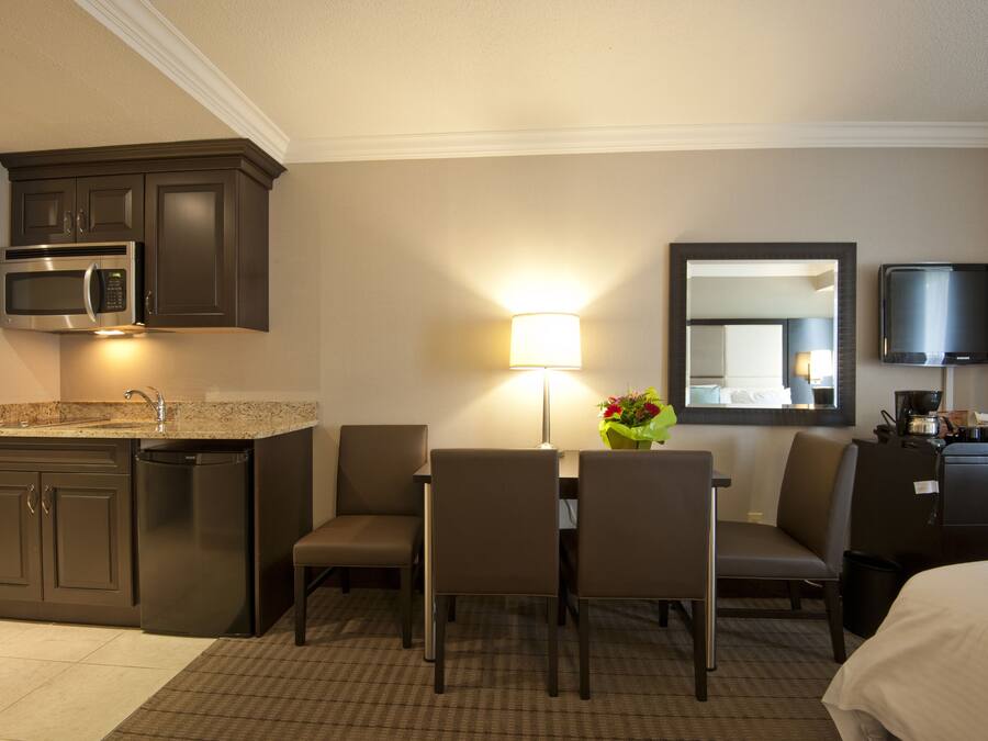 A hotel room with a kitchenette, a dining area with chairs and a table, a lamp, a mirror, and a TV.