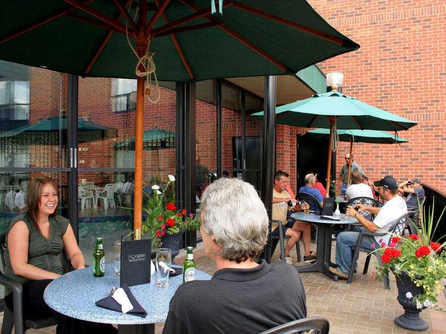 An outdoor patio area with round tables and green umbrellas, brick walls, and a pool in the background.