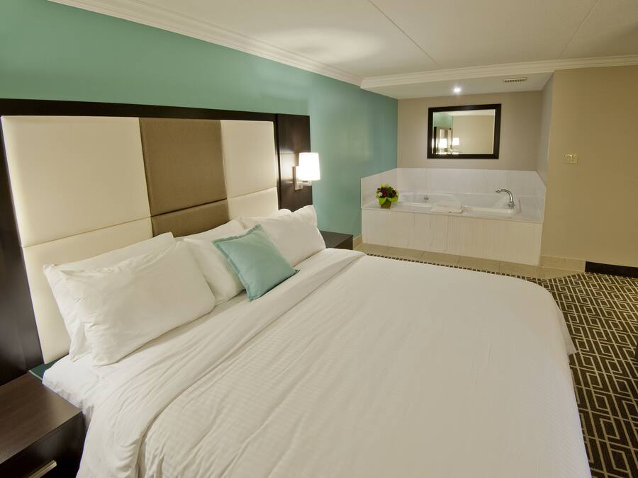 A large bed with white bedding and a teal pillow, a nightstand, a wall-mounted lamp, a bathroom with a bathtub and sink, and a patterned floor.