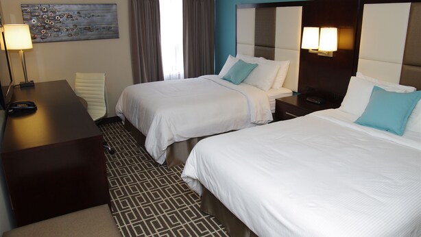 A hotel room with two beds, a desk, a lamp, and a painting on the wall. - Visitors Inn (Hamilton)