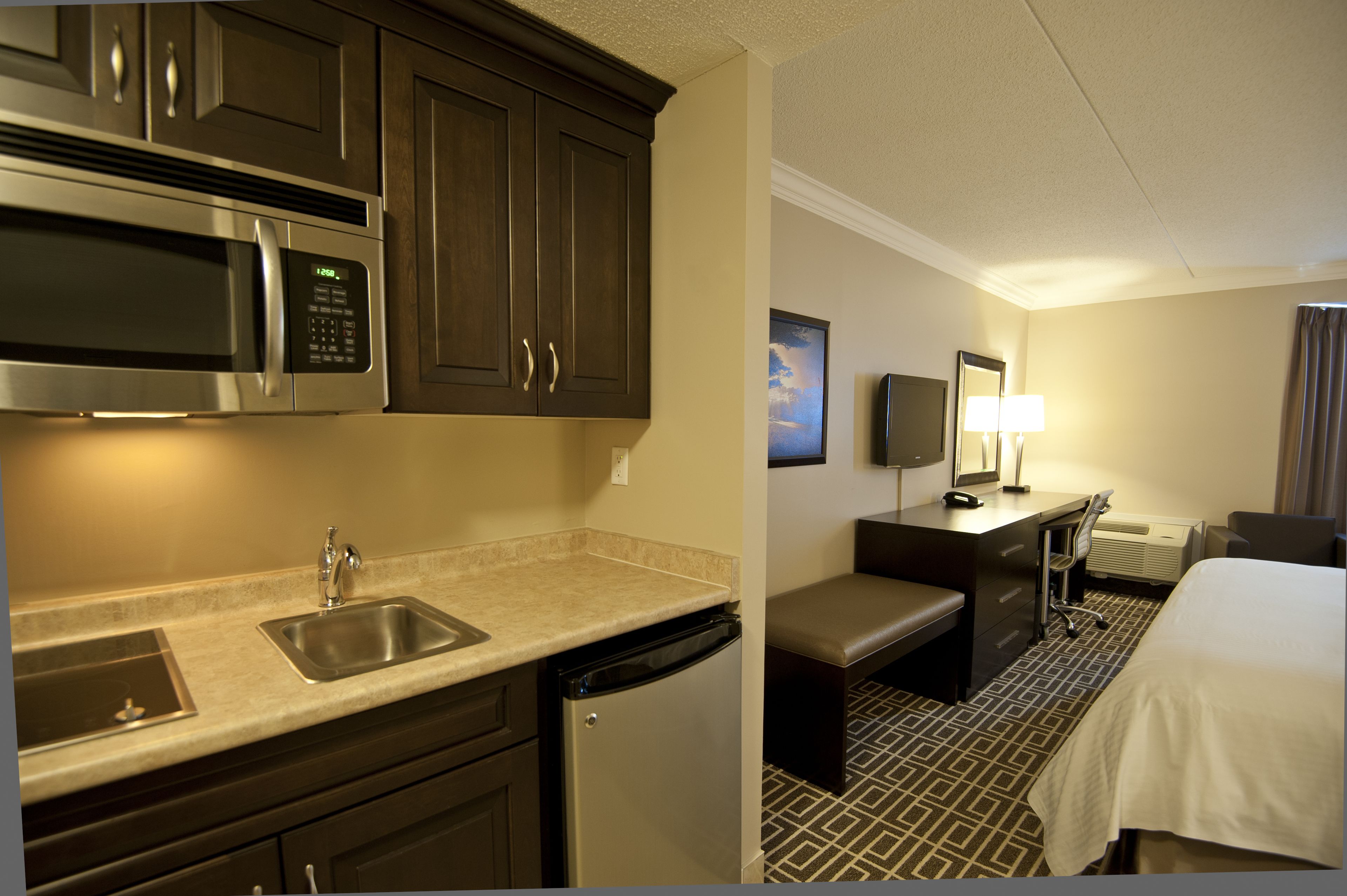 A compact hotel room with a kitchenette including a microwave, sink, and dishwasher. There is a small seating area with a bench and a desk with a chair.