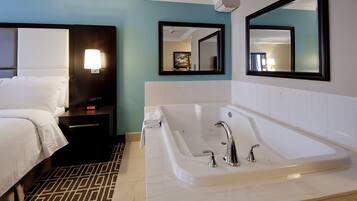 A hotel room with a large white bathtub, a bed with white linens, a nightstand with a lamp, and two wall-mounted mirrors.