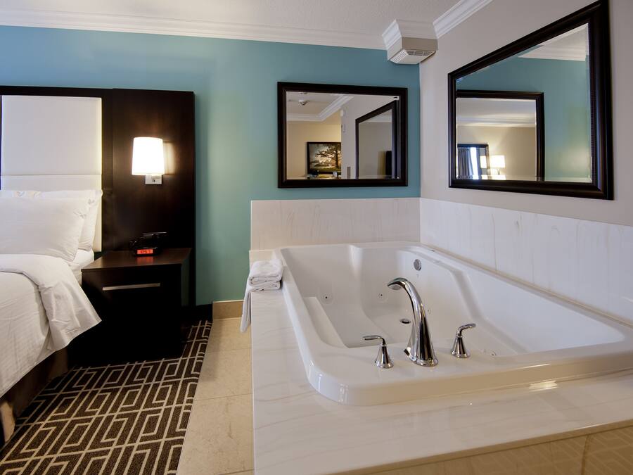 A hotel room with a large white bathtub, a bed with white linens, a nightstand with a lamp, and two wall-mounted mirrors.