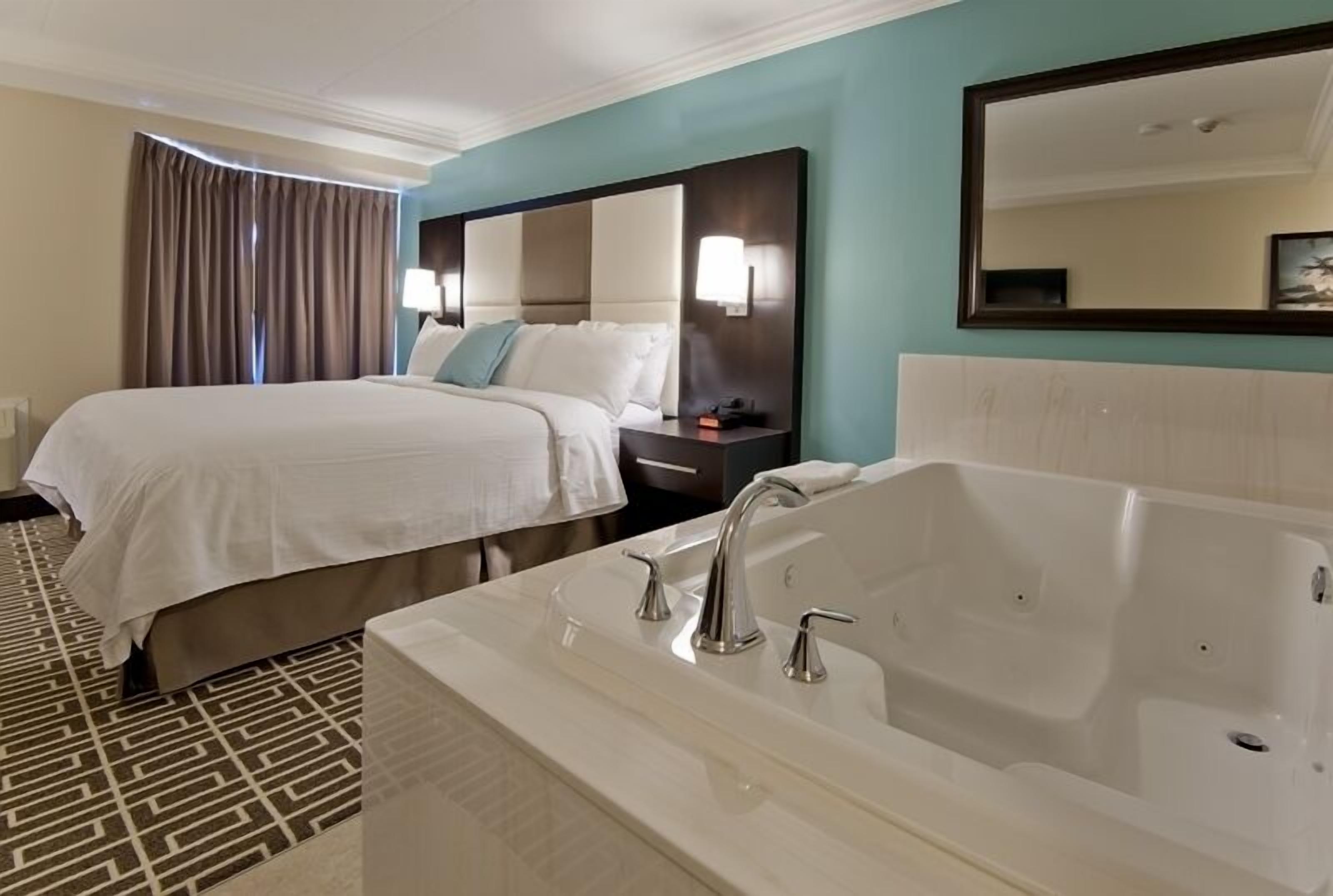 Quarto de hotel com uma cama grande, área de estar separada, banheira e um espelho amplo.