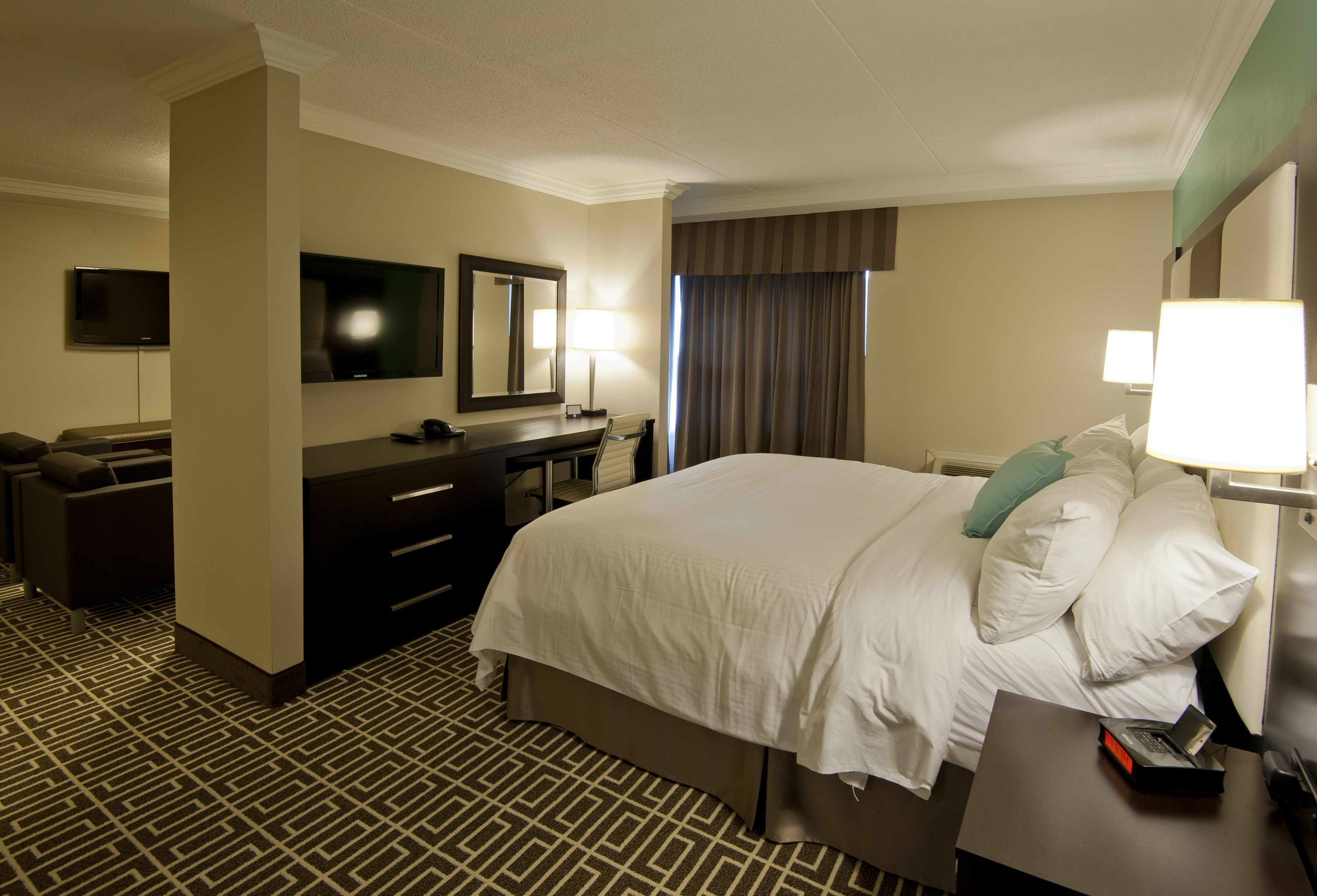Une chambre d  ’hôtel avec un grand lit, un bureau avec une chaise, un téléviseur à écran plat, un miroir, une lampe et une moquette à motifs.