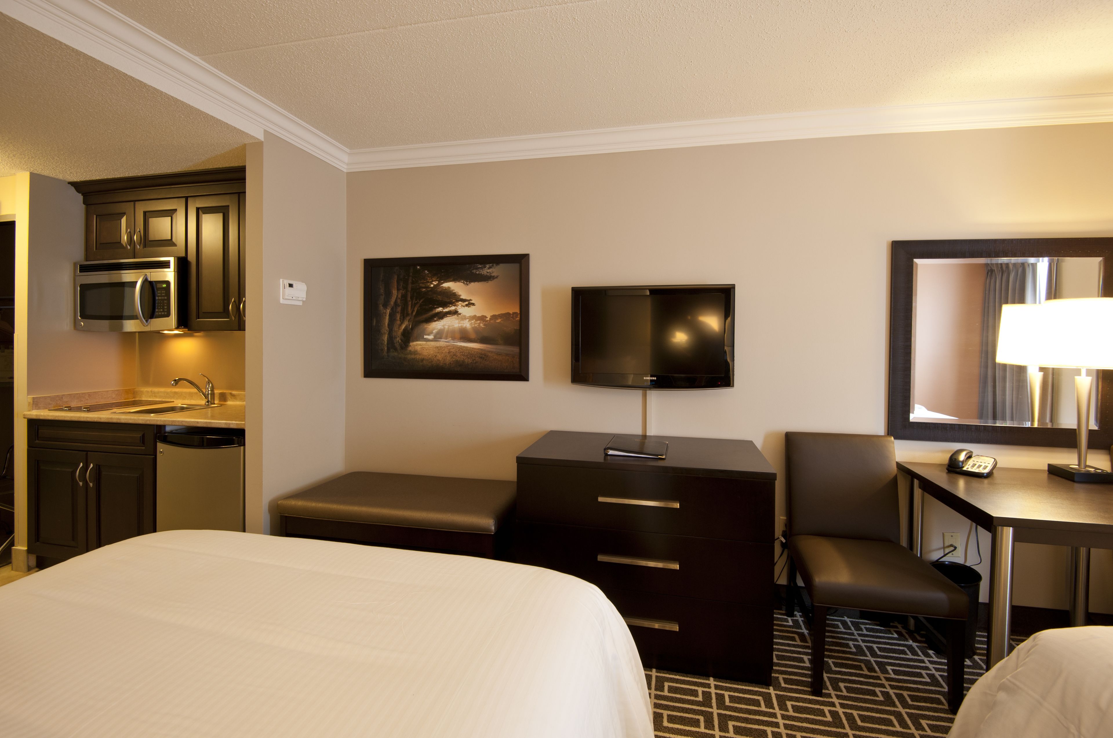 Habitación de hotel con cama, escritorio con lámpara, televisor, silla, mesita pequeña, microondas y lavamanos.