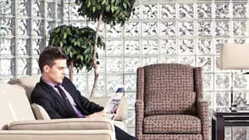 A man sitting on a beige sofa in a room with patterned wallpaper, a patterned armchair, and a small table with a lamp.