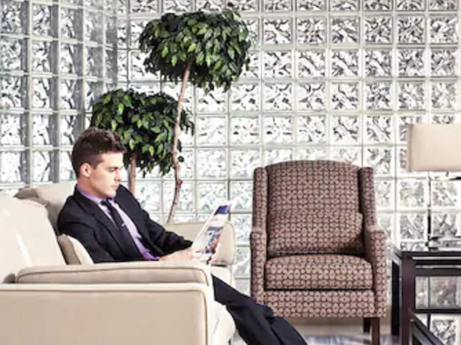 A man sitting on a beige sofa in a room with patterned wallpaper, a patterned armchair, and a small table with a lamp.