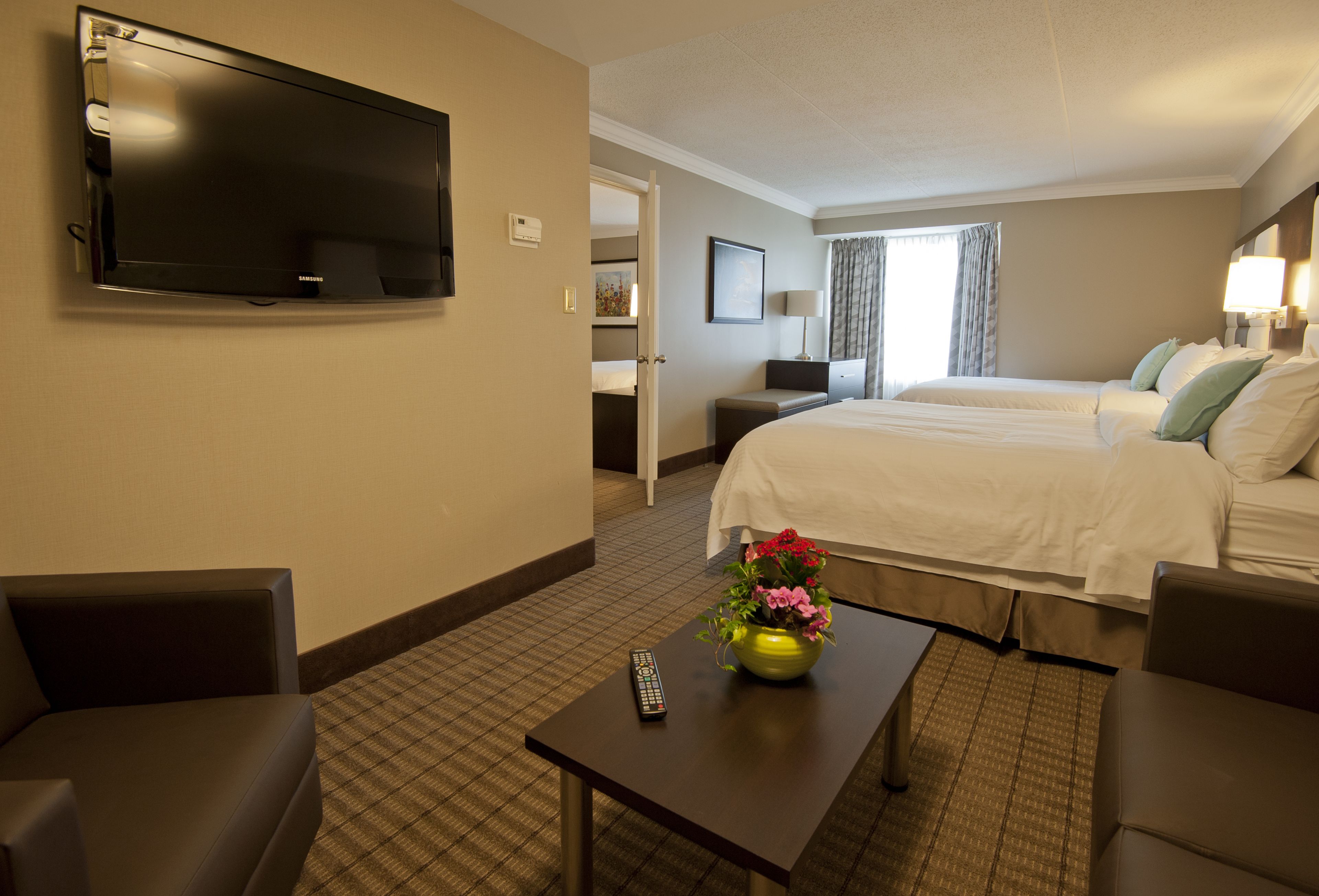 A hotel room with a flat-screen TV mounted on the wall, a bed with white linens, a small table with a vase of flowers, and a remote control on the table.