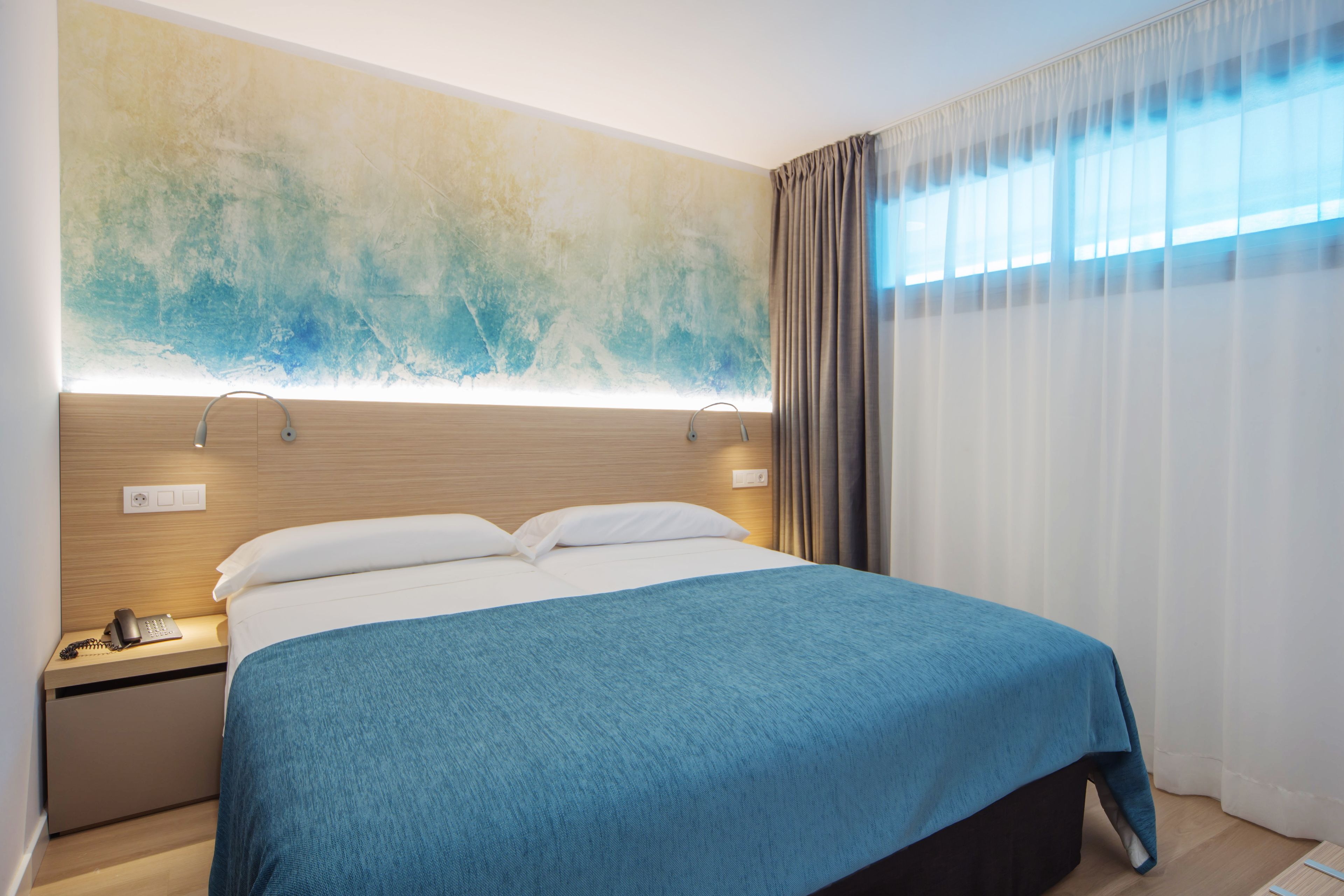 Habitación de hotel moderna con una cama grande, una mesita de noche con teléfono y una lámpara fijada a la pared.
