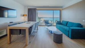 Une chambre d’hôtel moderne dotée d’une grande fenêtre donnant sur la mer, d’un coin repas avec une table et des chaises, d’un canapé et d’un téléviseur à écran plat fixé au mur.