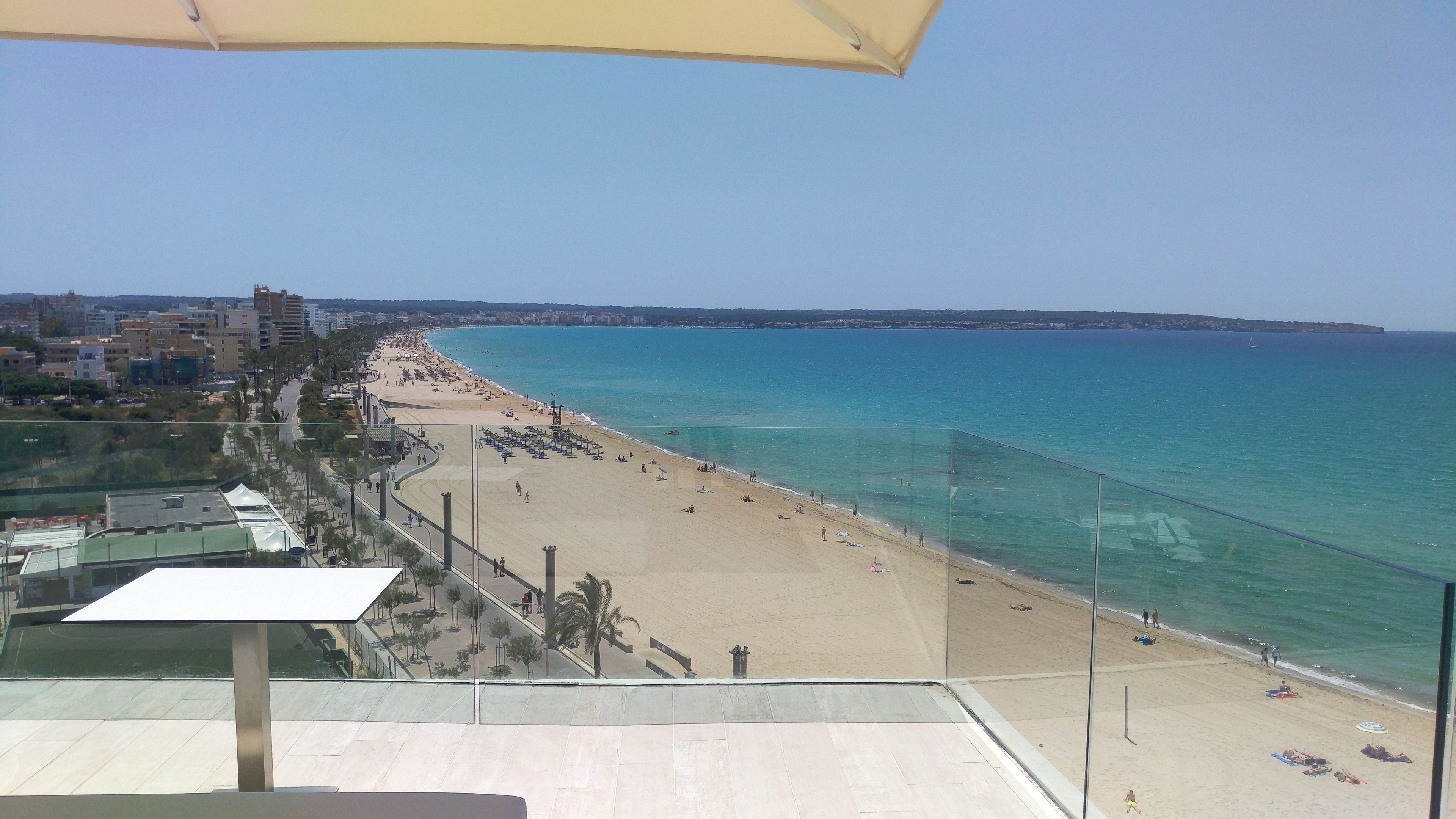 Vista de la playa desde un balcón, cielo despejado, arena dorada y mar tranquilo.