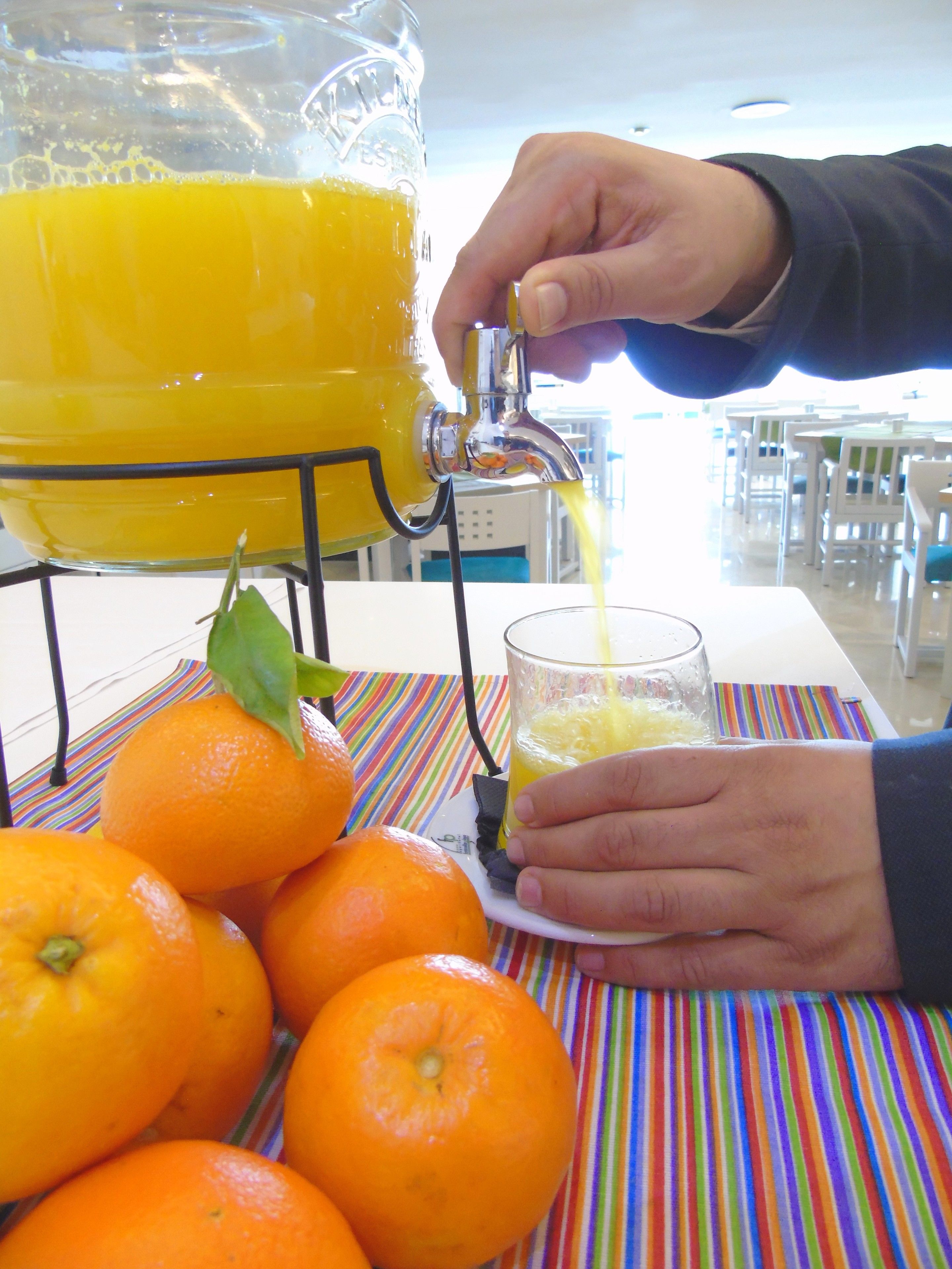 Une personne verse du jus d’orange d’une grande carafe dans un verre. On peut voir des oranges et une nappe à rayures à l’arrière-plan.