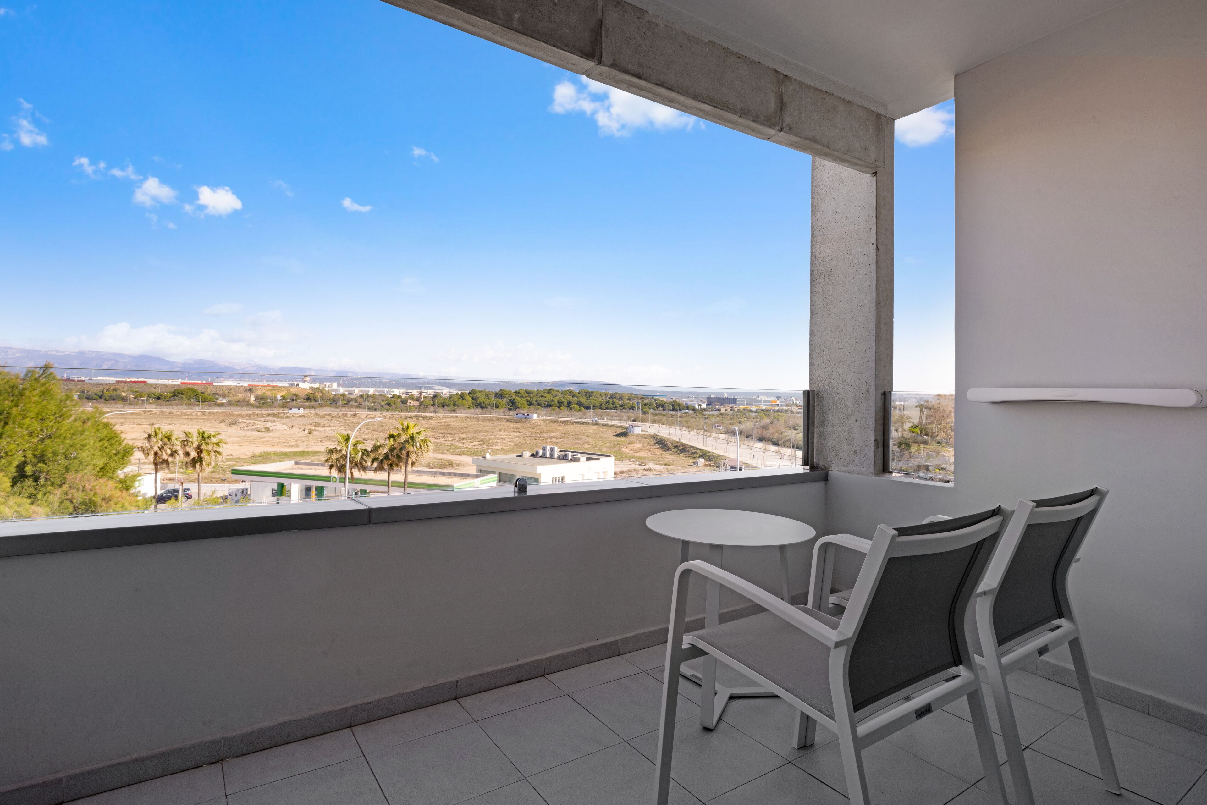A balcony with two white chairs, a small round table, and a view of a distant landscape with buildings and trees.