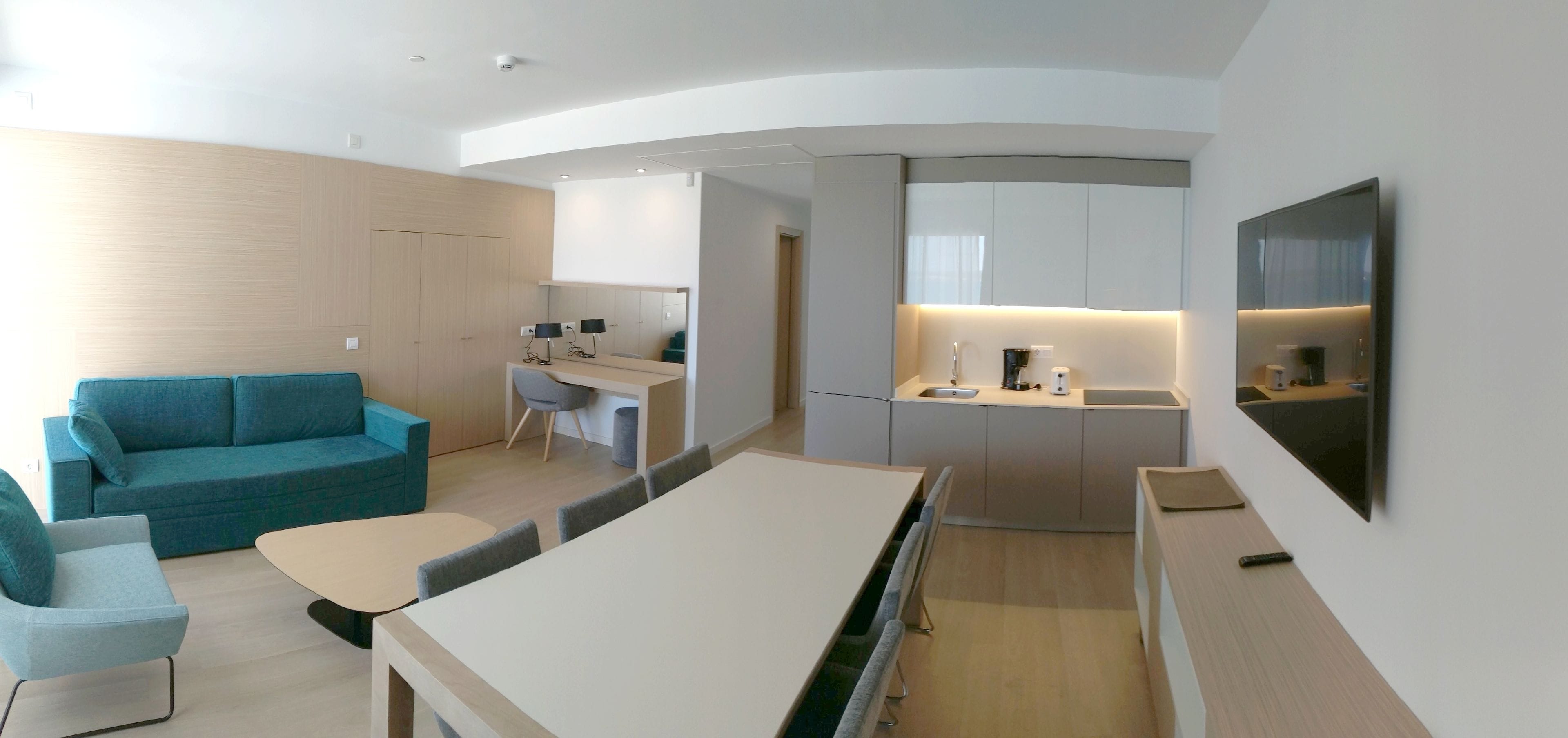 A modern, compact living space with a sofa, a dining table, a kitchen area with a sink and countertop, and a TV mounted on the wall.