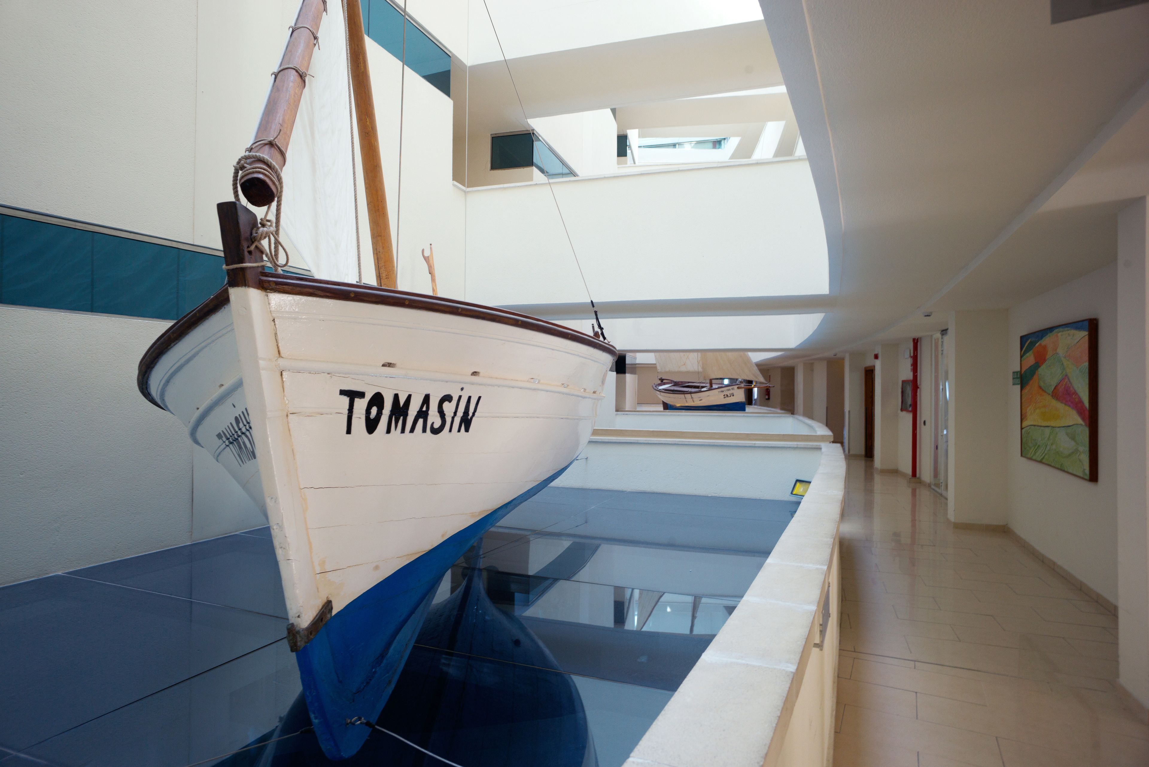 Una barca anomenada 'TOMASIN' en un espai interior modern amb un sostre de vidre, un passadís i un quadre a la paret.