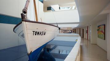 Een boot genaamd 'TOMASIN' in een moderne, overdekte ruimte met een glazen vloer, een gang en een schilderij aan de muur.