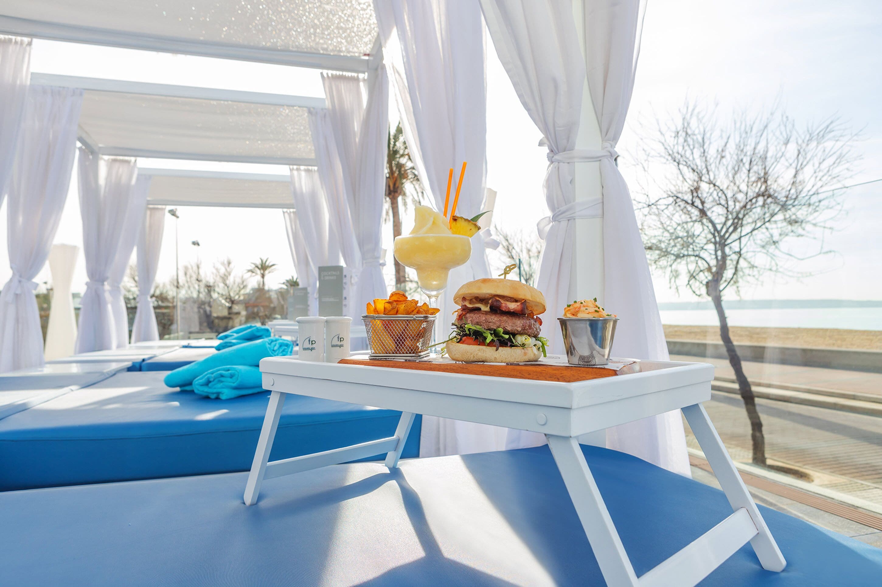 Une pièce baignée de lumière, avec une piscine, une table sur laquelle on trouve un hamburger, une boisson et des frites.