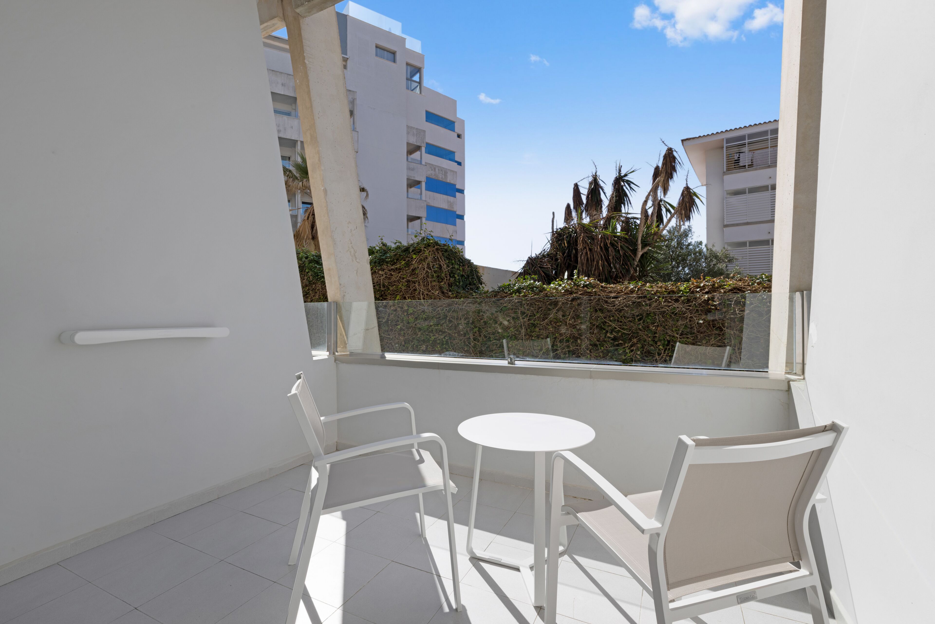 Un balcone con sedie bianche e un tavolino, affaccia su una veduta cittadina con edifici e palme.
