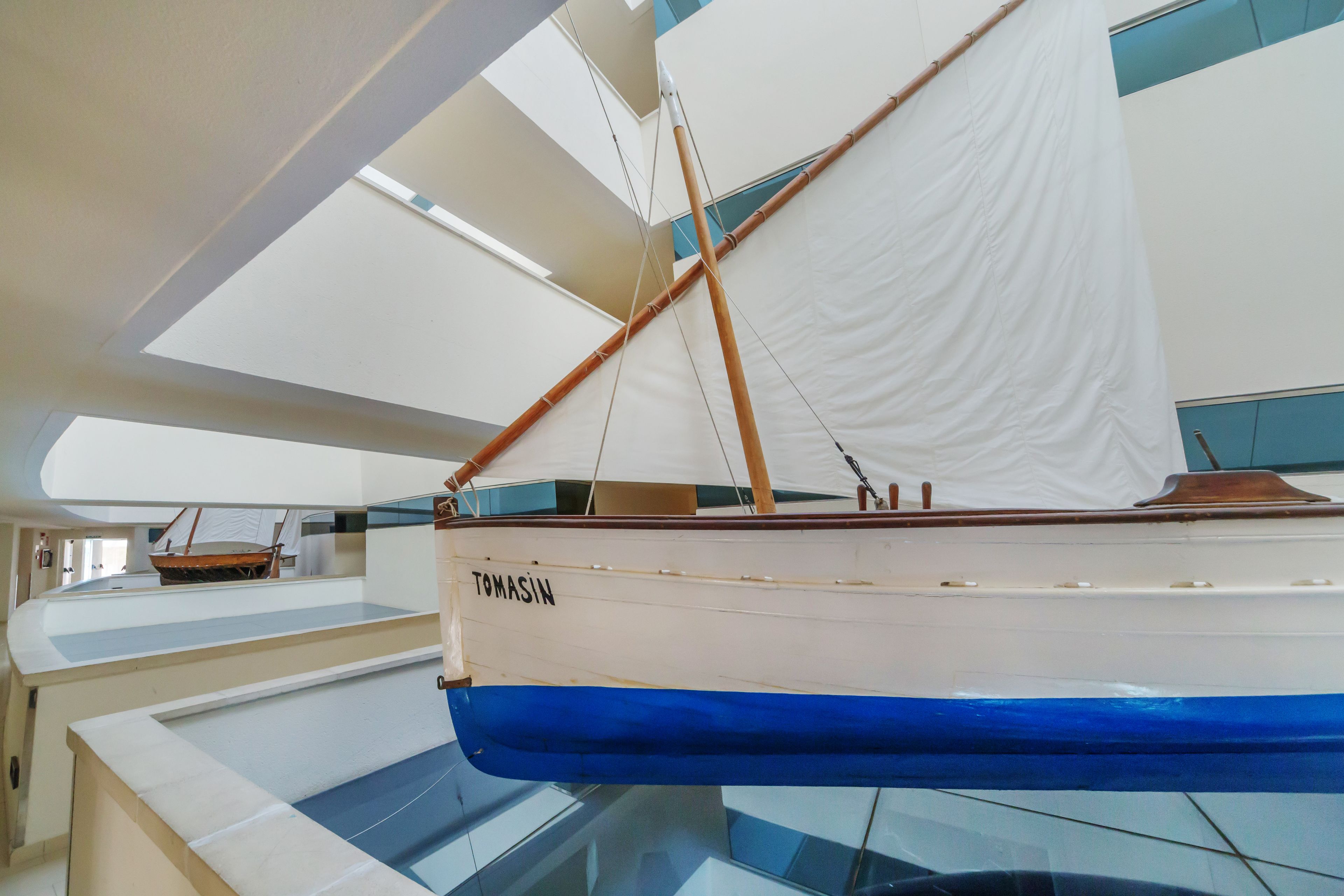 A boat named TOMASIN displayed in a museum or exhibit space with glass flooring and a high ceiling.