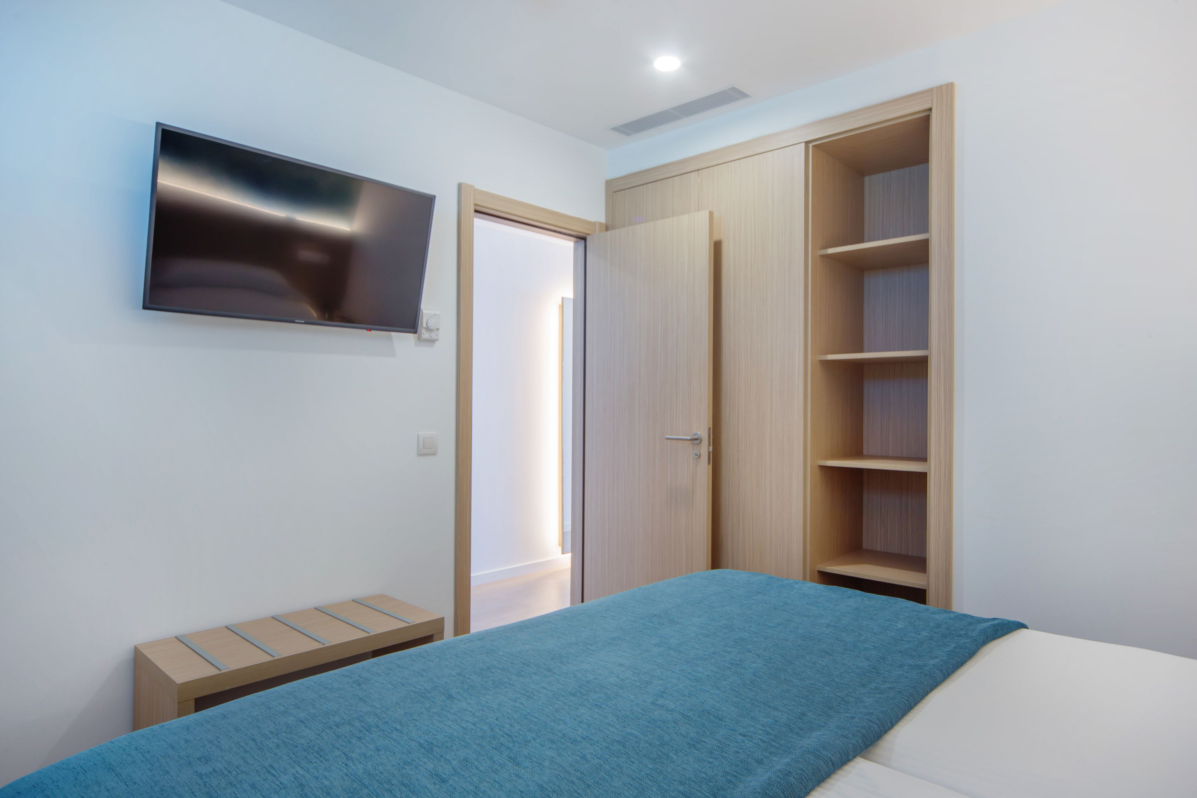 A bedroom with a bed, a TV mounted on the wall, a wooden wardrobe, and a small bench.
