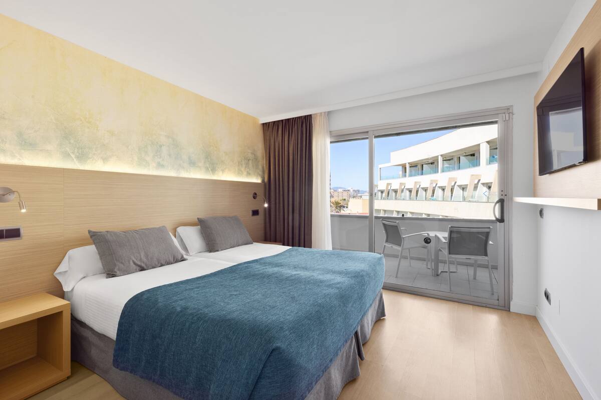 Habitación de hotel moderna con una cama grande, un balcón con mobiliario exterior y vistas a la ciudad.