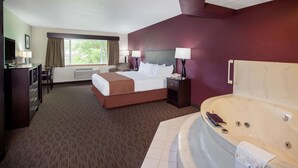 Habitación de hotel con una cama grande, una zona de estar con escritorio y silla, una bañera grande y una ventana con cortinas.
