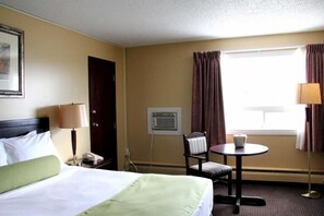 A hotel room with a bed, a desk, a chair, a window with curtains, and a wall-mounted air conditioning unit.