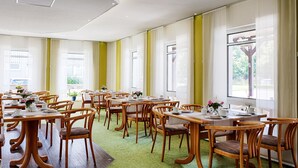 Un comedor con mesas y sillas de madera, ventanas amplias con cortinas y una alfombra verde.
