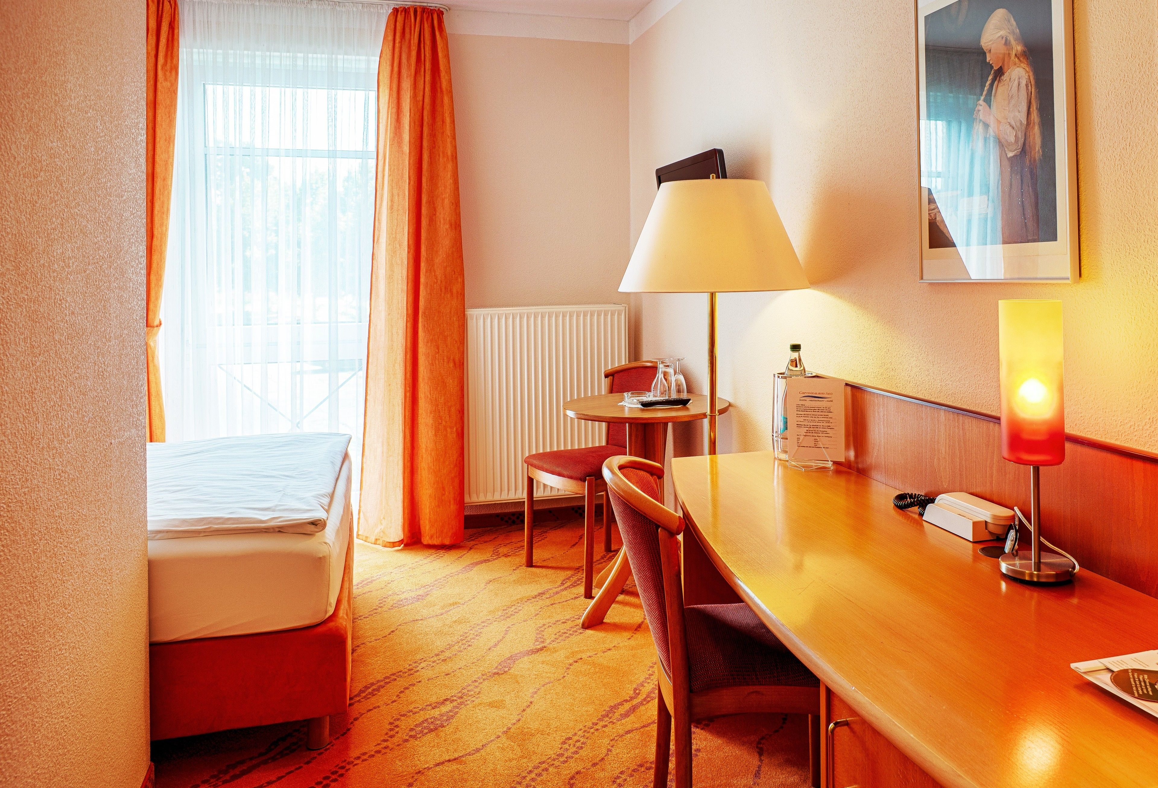 A hotel room with a bed, a desk with a lamp, a chair, a small table, and a framed picture on the wall.