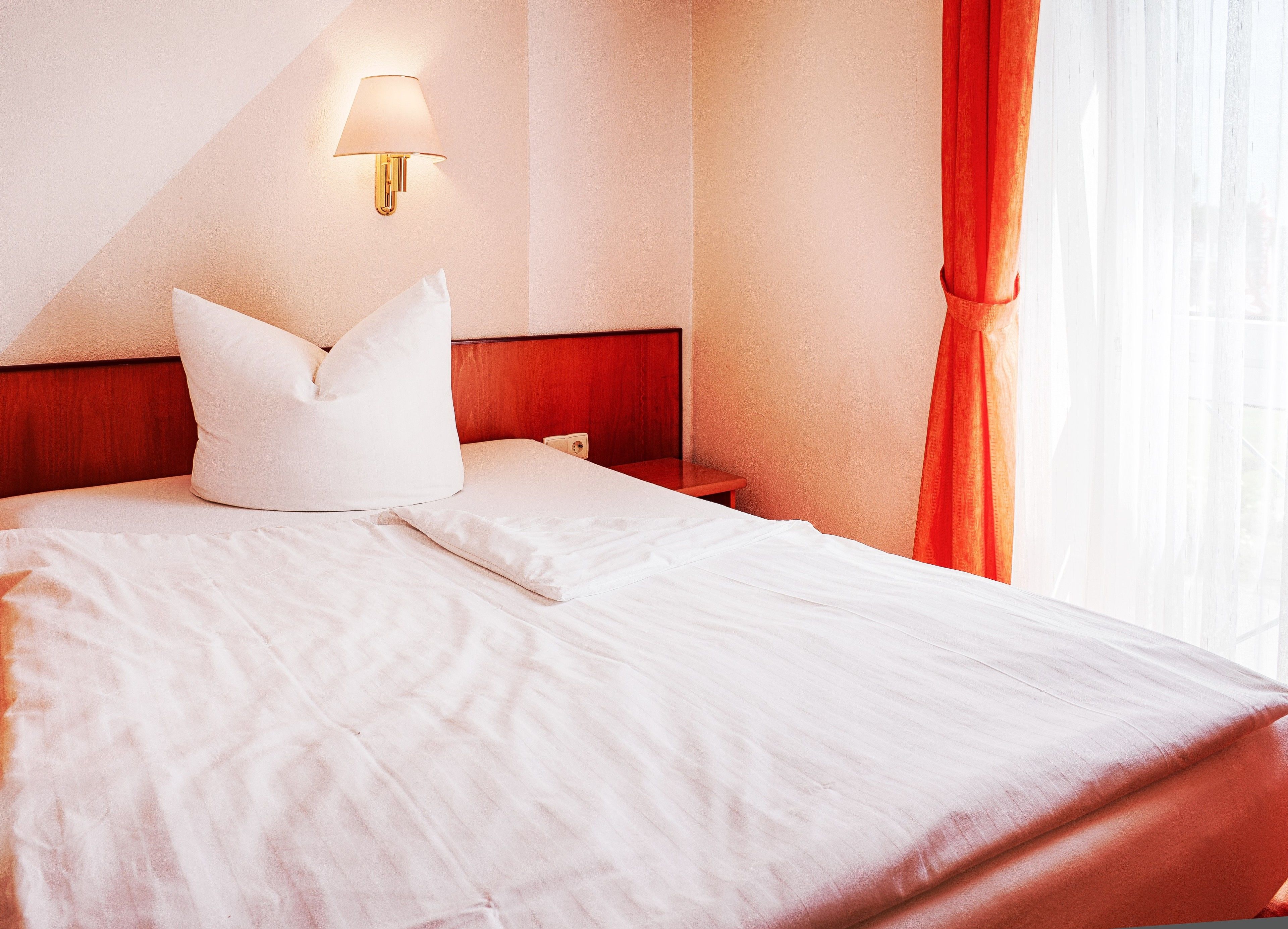 A neatly made bed with white linens, a wooden headboard, a wall-mounted lamp, and a window with orange curtains.