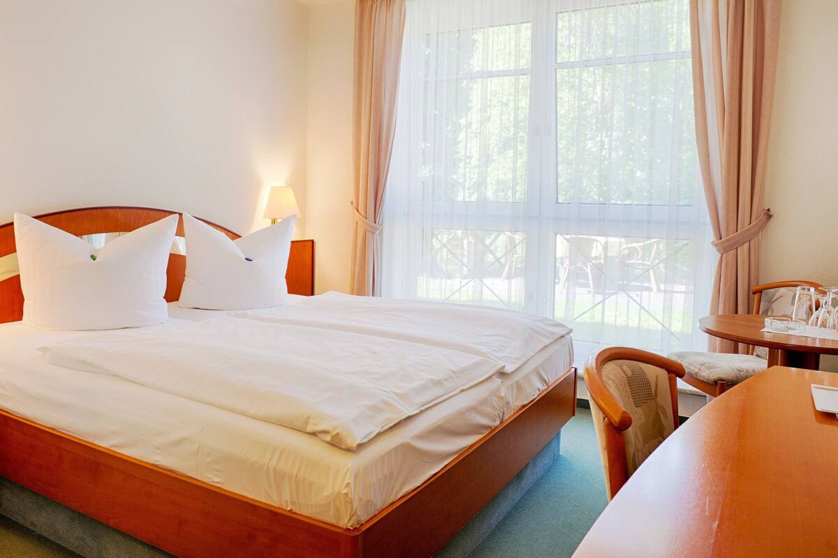 A hotel room with a large bed, a wooden headboard, a bedside table with a lamp, a small table with a chair, and a window with curtains.
