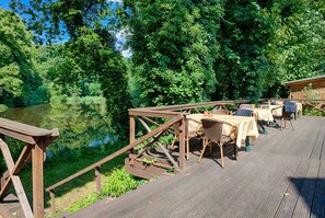A wooden deck with tables and chairs overlooking a pond, surrounded by dense greenery. - Hotel Carmina am See (Karnin)
