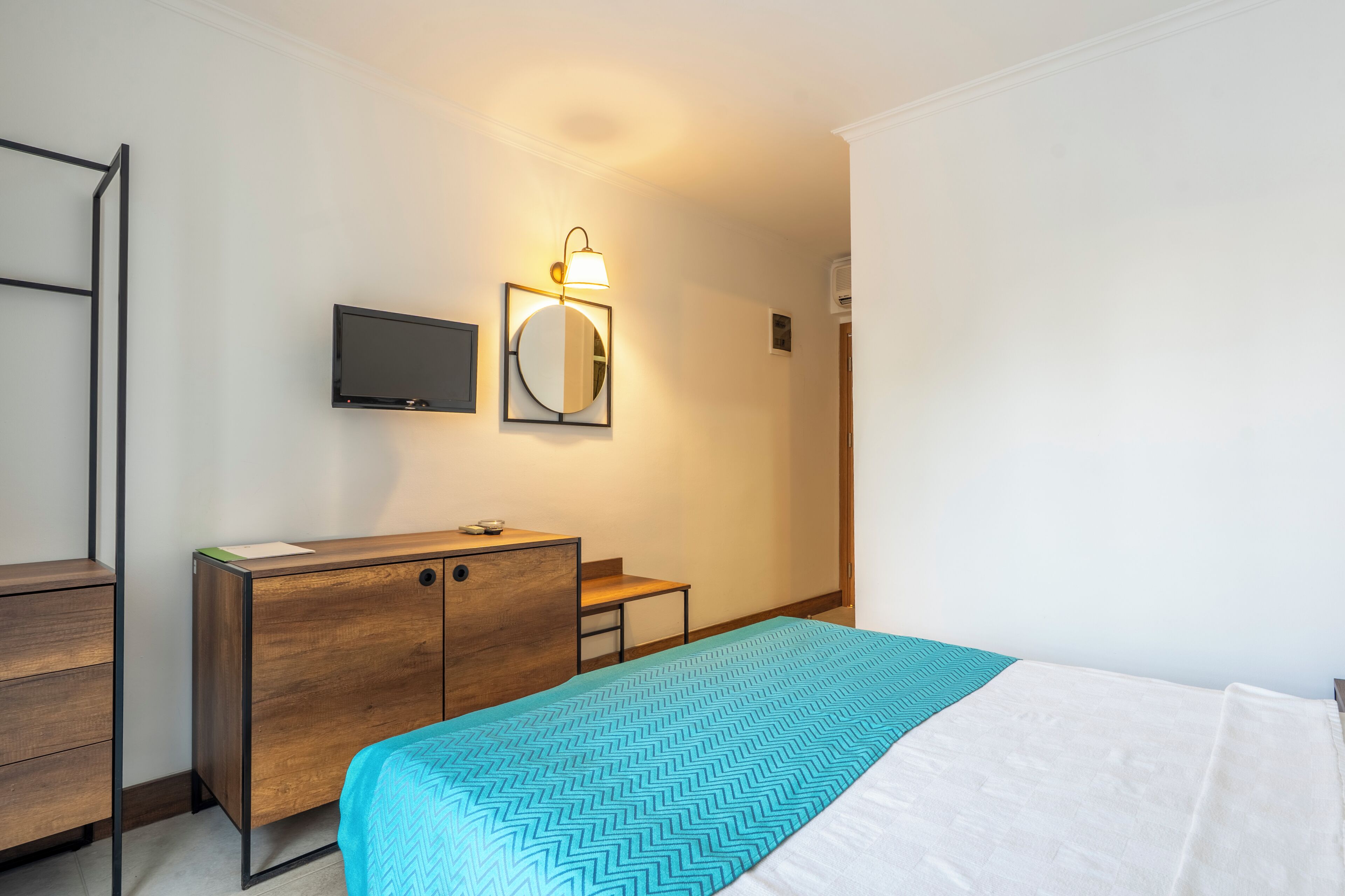 A hotel room with a bed, a wooden dresser, a desk, a television, and a wall-mounted mirror.