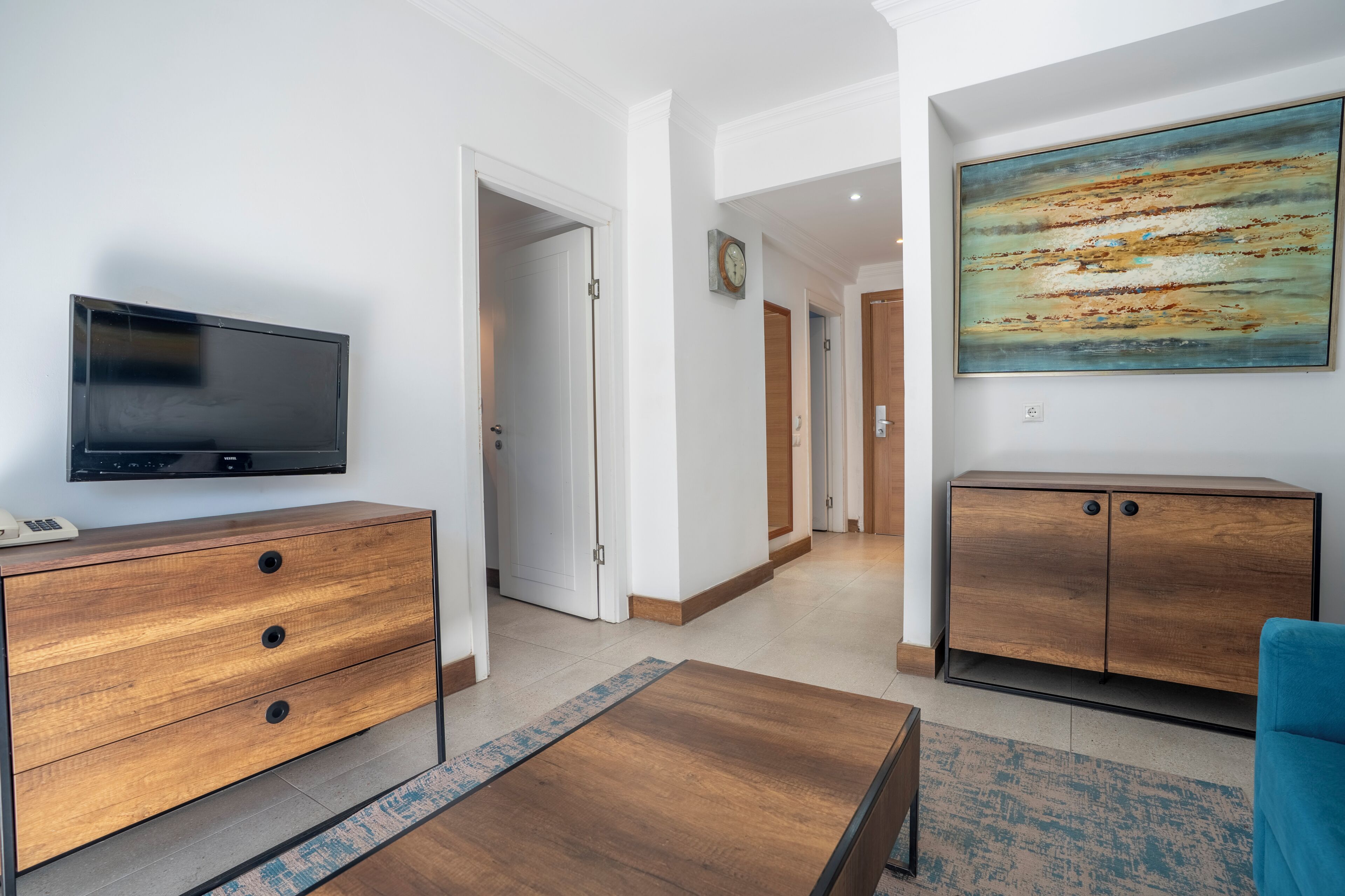 Een moderne woonkamer met een flatscreen televisie, houten meubels, een schilderij en een gang die naar andere kamers leidt.