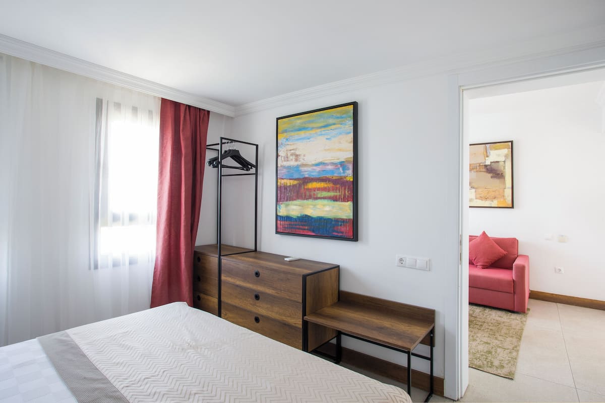 A bedroom with a bed, a wooden dresser, a desk, a red sofa, and two paintings on the wall.