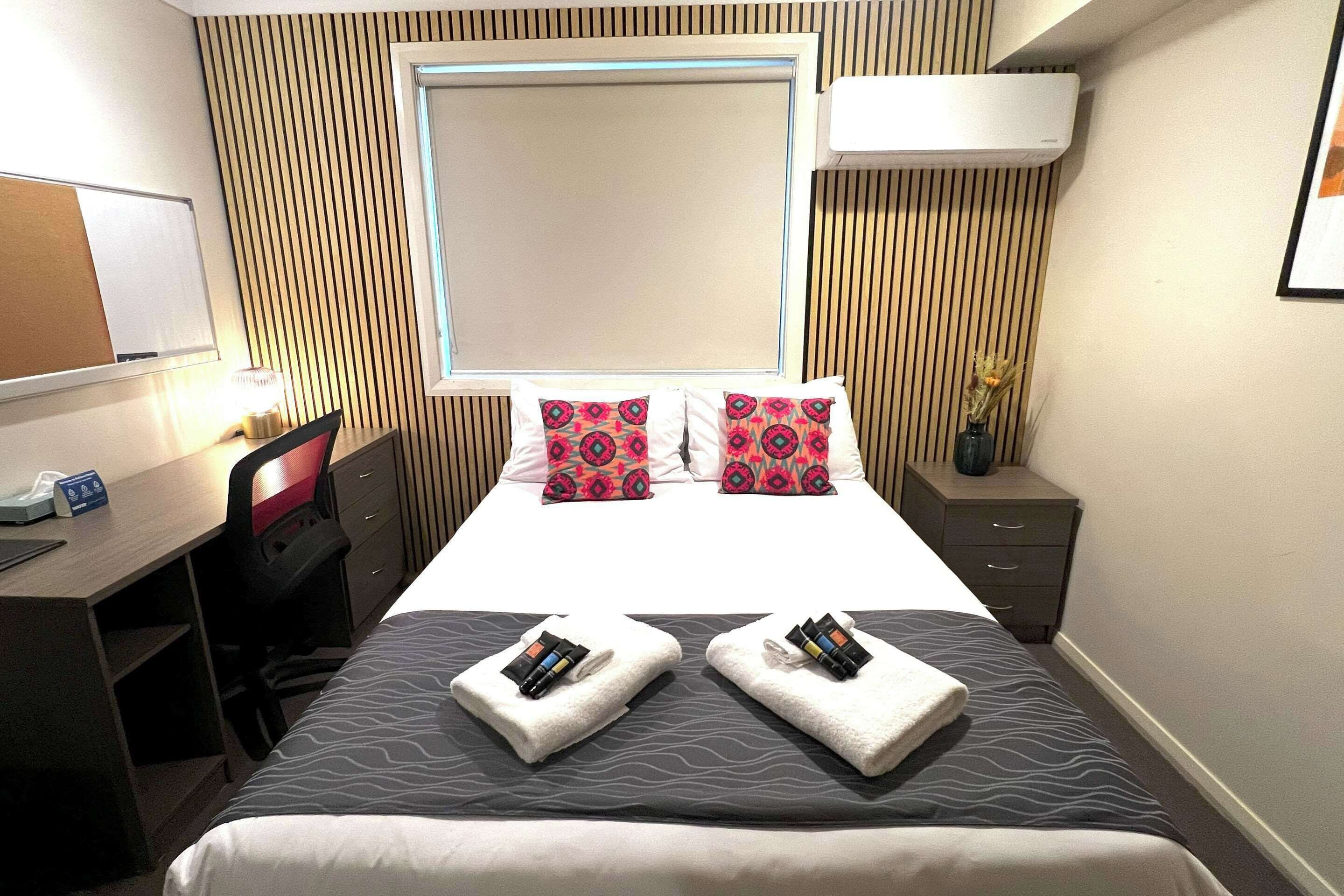 Une chambre d’hôtel comprenant un lit, deux oreillers, un bureau, une chaise, une table de chevet, une lampe et une fenêtre.