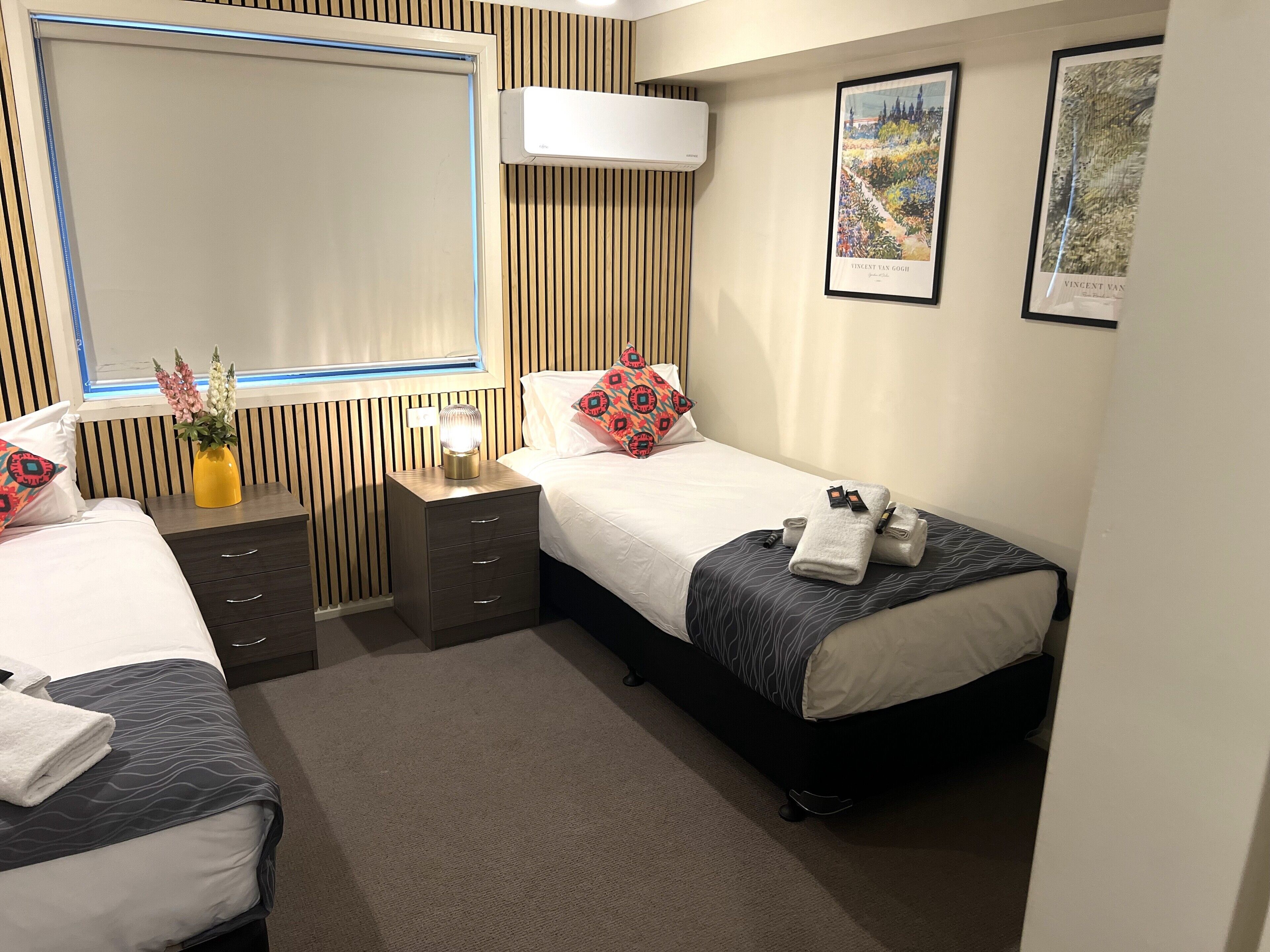 Habitación de hotel con dos camas, una mesita de noche con lámpara, aire acondicionado instalado en la pared y dos cuadros enmarcados.